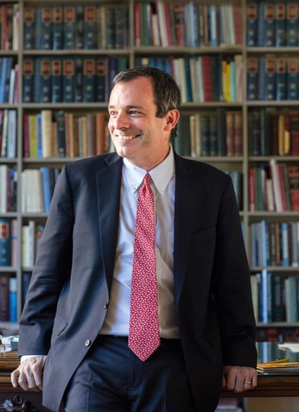 Thomas Manning in a suit, smiling and leaning on a desk while looking to his right