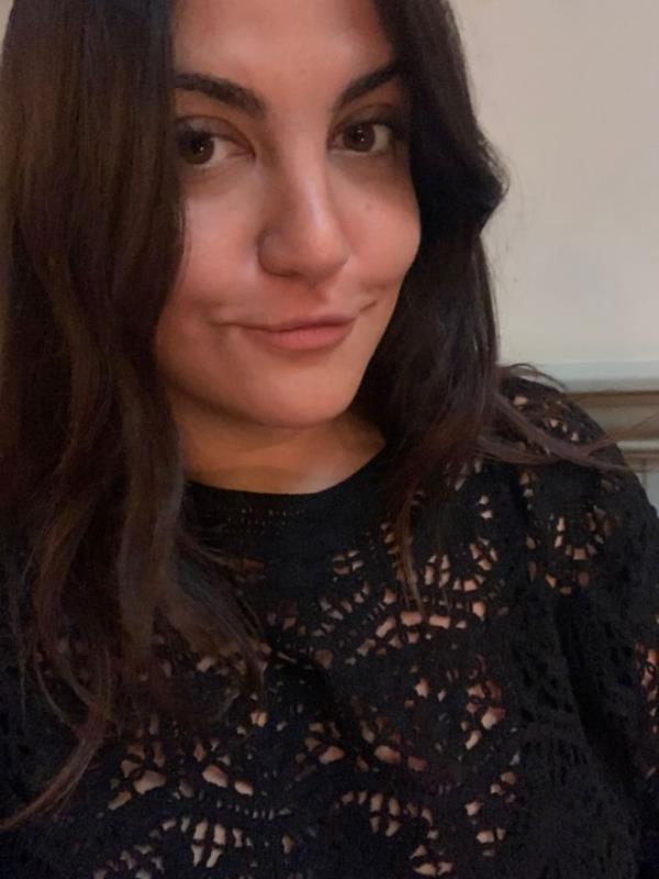 A selfie of Almara smiling to camera, with long dark hair, wearing a black crocheted top.
