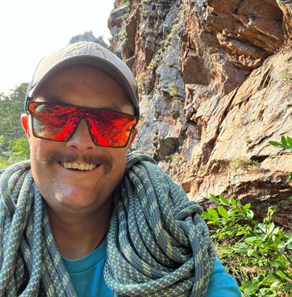 Levi Wilkins smiles with a rock climbing rope over his shoulders and a cliff in the background. 