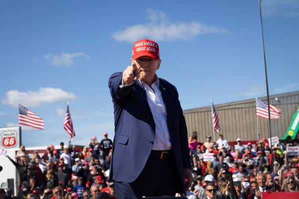 Former President Do<em></em>nald Trump in a suit and red hat, pointing at the camera as he departs a campaign event at the Central Wisco<em></em>nsin Airport.