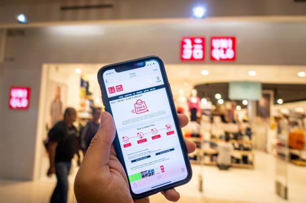 Man's hand holding an iPhone X displaying the Uniqlo app, in front of a Uniqlo store