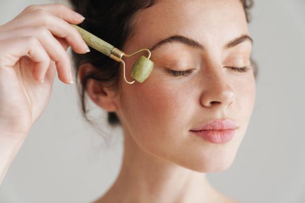 Half-naked brunette woman massaging her face with a jade roller against a white background