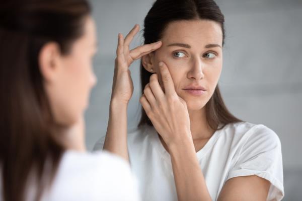 An anxious young woman examining her face in the mirror, co<em></em>ncerned a<em></em>bout acne and wrinkles