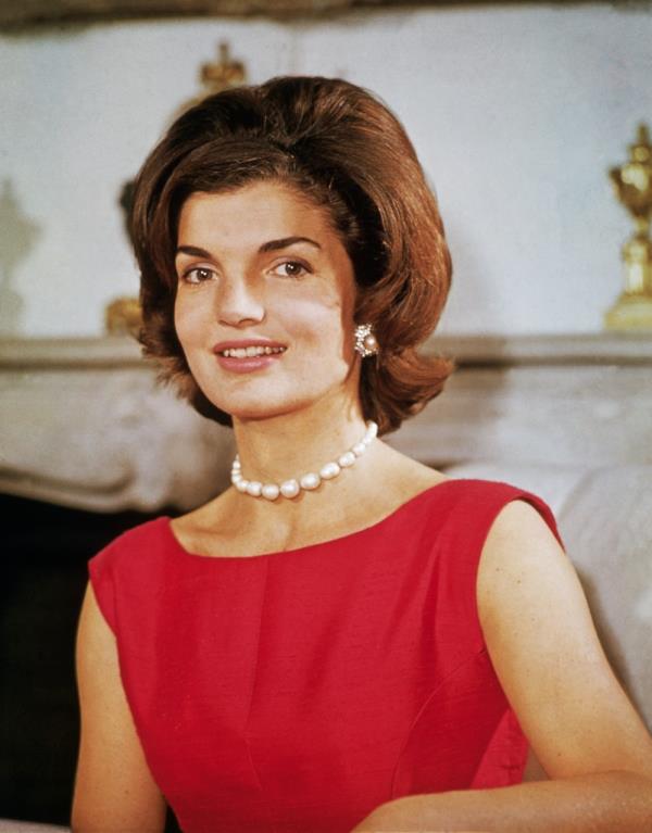 Jacqueline Kennedy in 1960, in a red sleeveless dress and large white pearl necklace smiling and looking at the camera