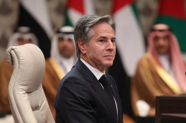 A close up of U.S. Secretary of State Antony Bl<em></em>inken seated in a chair, with blurred images of Arab leaders wearing headdresses and robes in the background.