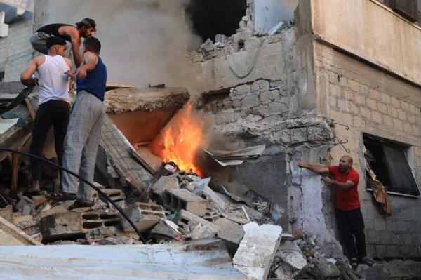 Three Palestinian men stand atop the rubble of a destroyed building, with a fourth gesturing at them from across the pile and a fire burning between them.