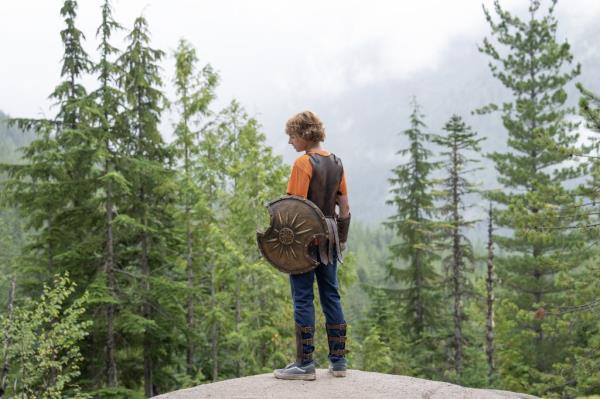 Walker Scobell standing on a rock surrounded by tall pine trees. 