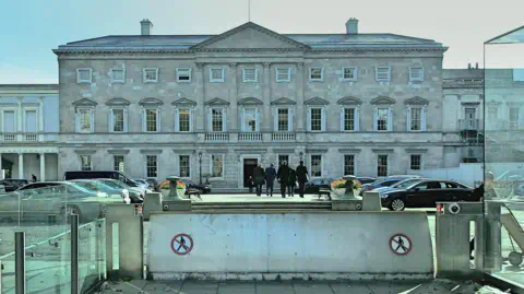 Getty Images Government Buildings on Kildare Street in Dublin with a security barrier raised. Some people in suits are walking into the building. There are cars parked outside. There's a sign on the barrier showing a person walking in a circle with a red line crossed through it.